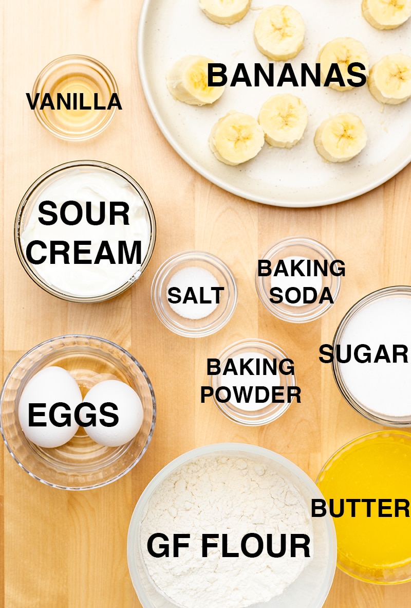 ingredients for gluten free banana bread including sliced bananas, vanilla, sour cream, salt, leaveners, eggs, white powder in bowls, melted butter, and white sugar in small bowls on blond wood table