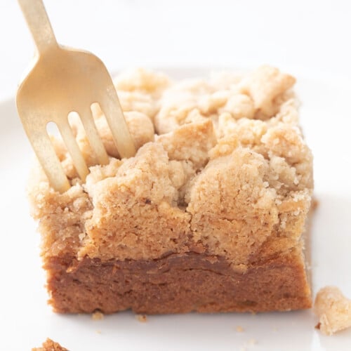 gold fork piercing light brown crumble topping of gluten free coffee cake slice on small white plate