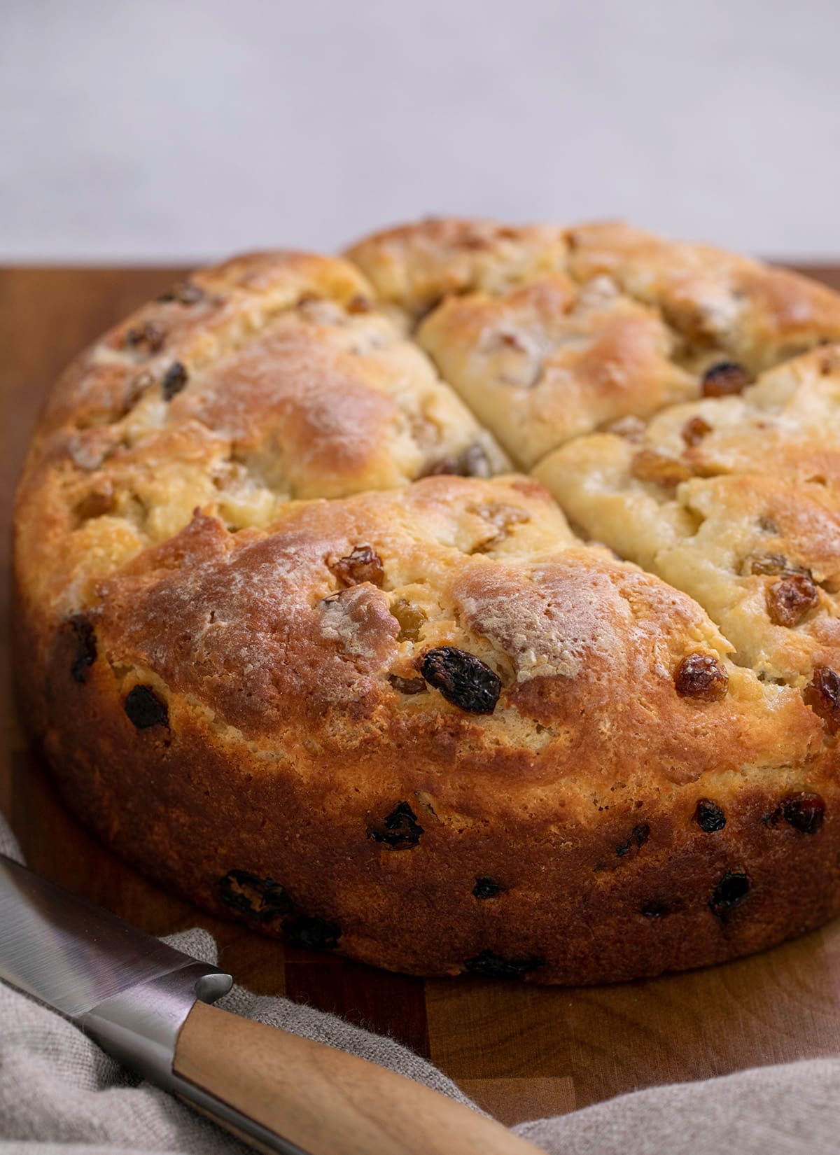 Whole brown flat round loaf of Irish soda bread on on brown cutting board with bread knife and tan cloth