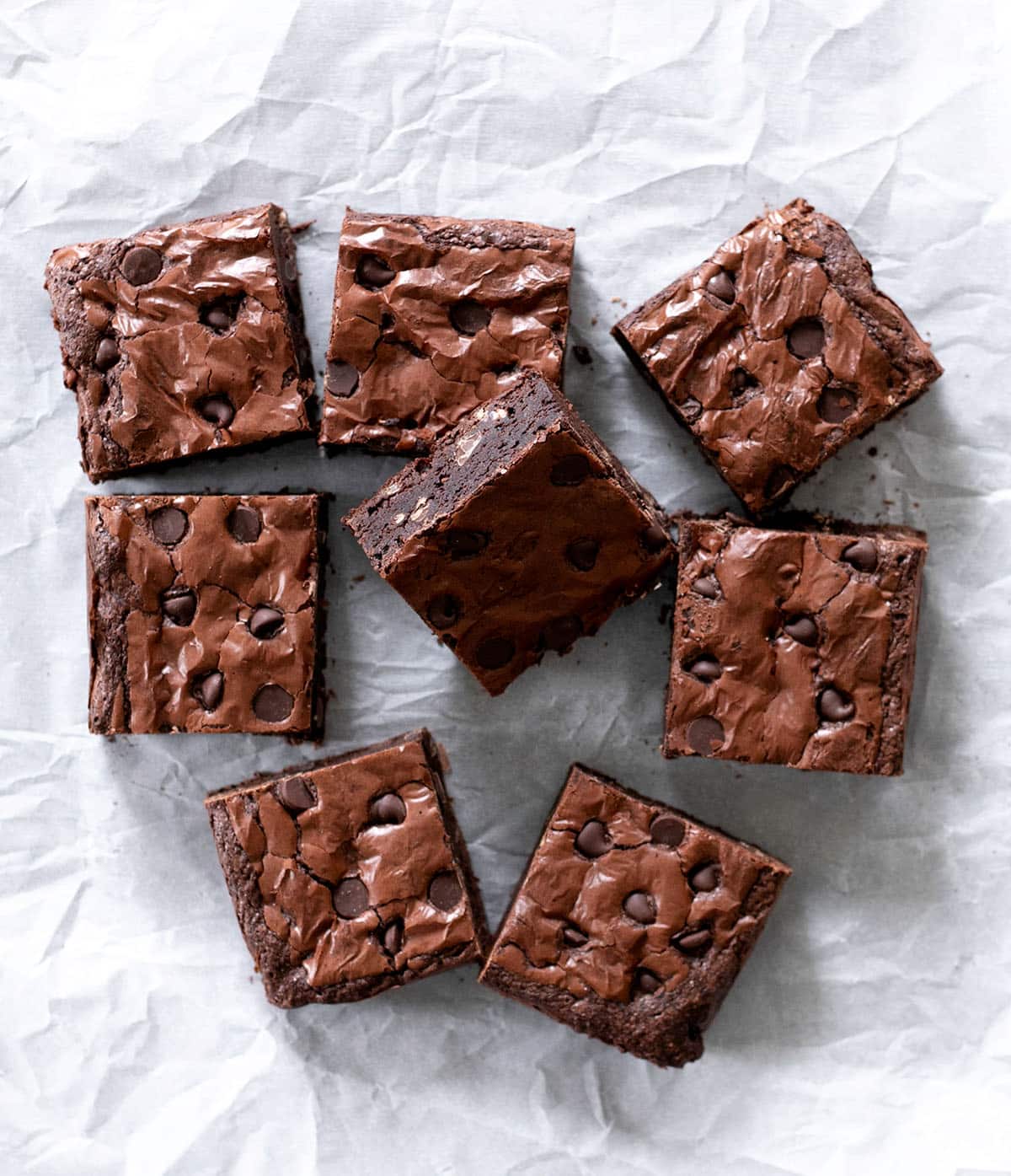 Overhead image of 8 brown bar squares with chocolate chips on white crinkly paper.