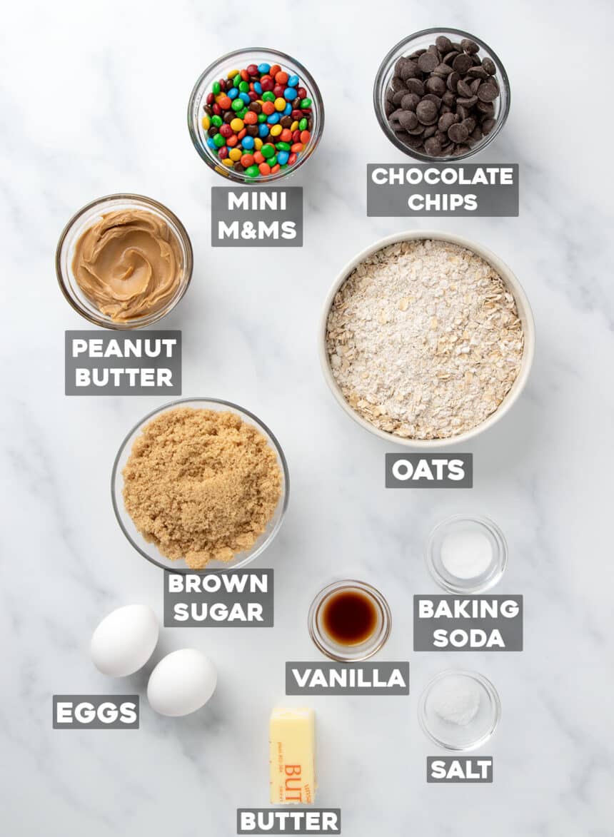 overhead image of gluten free monster cookies ingredients in small bowls with names of ingredients, all on a marble surface