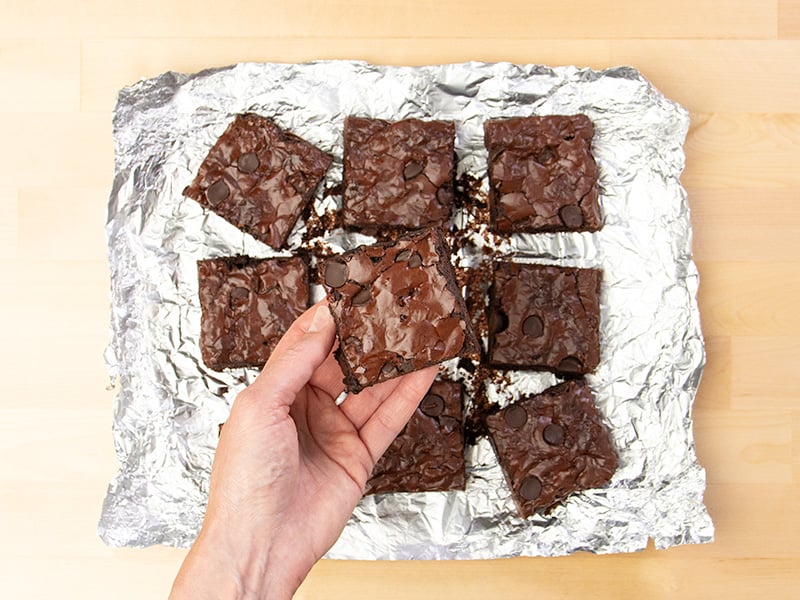 Hand holding gluten free brownie from center of 9 brownies on aluminum foil