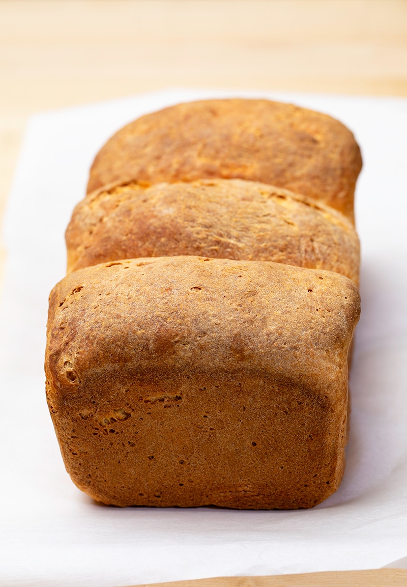 Whole loaf of bread with brown crust made of 3 joined parts on white paper on blonde wood table