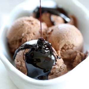 4 scoops of chocolate ice cream in white dish with dark chocolate syrup pouring on top