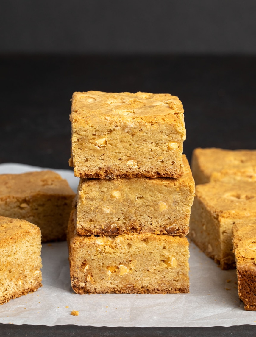 9 gluten free blondie squares, 3 in center of photo in a vertical stack, all on white parchment paper