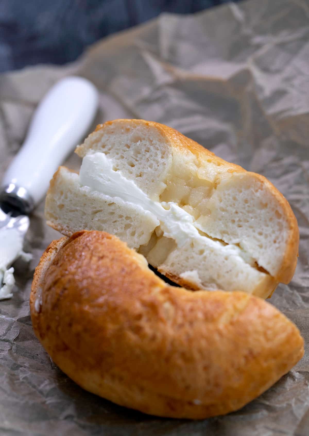 Baked light colored interior of brown crusted bagel sliced in half with cream cheese in center on brown paper