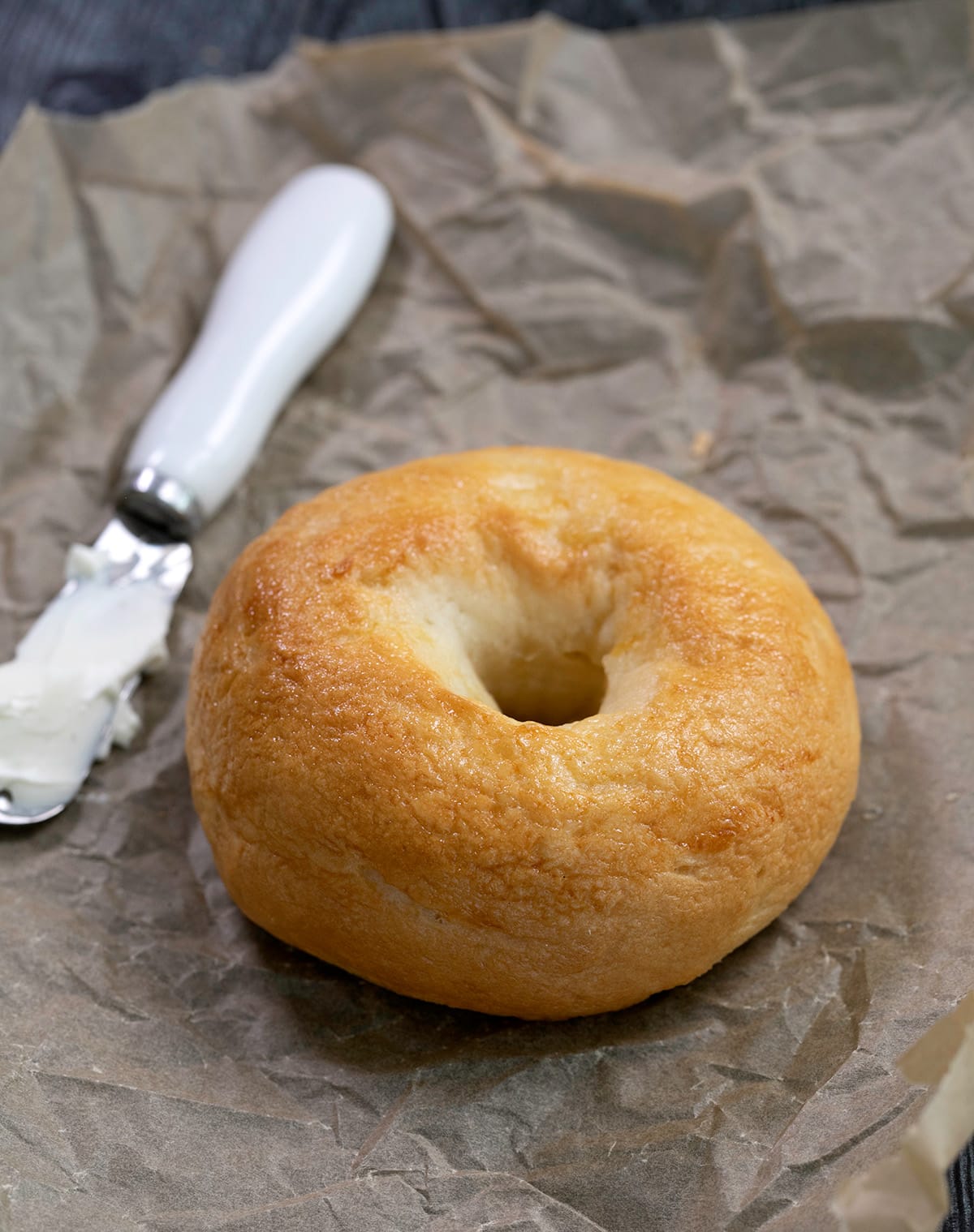 Light brown bagel on brown paper with a knife and cream cheese on a dish