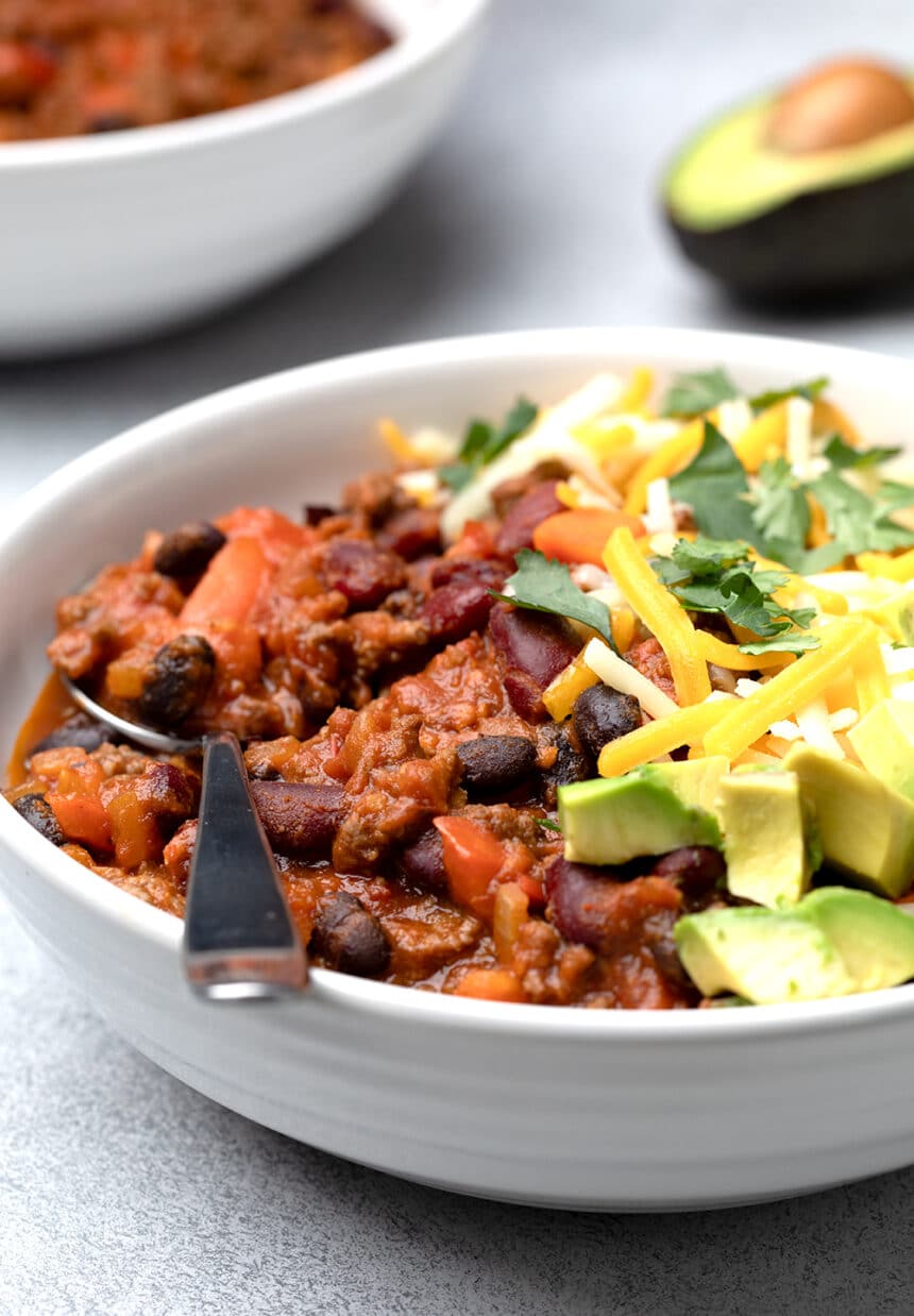 Red beef and bean gluten free chili in wide flat white bowl with spoon and toppings image from side