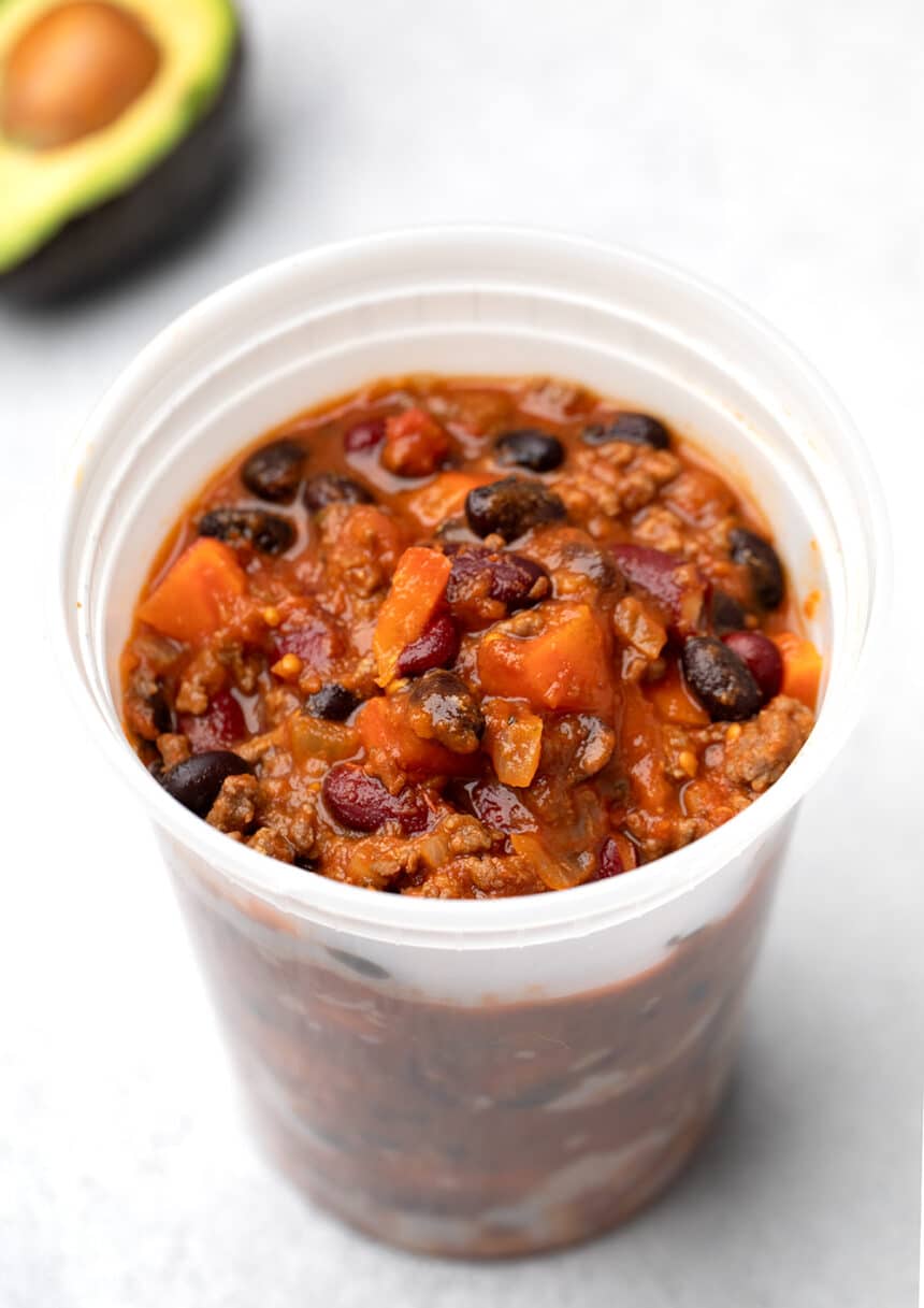 Red beef and bean and carrot chili in plastic deli container on white background with avocado half in background