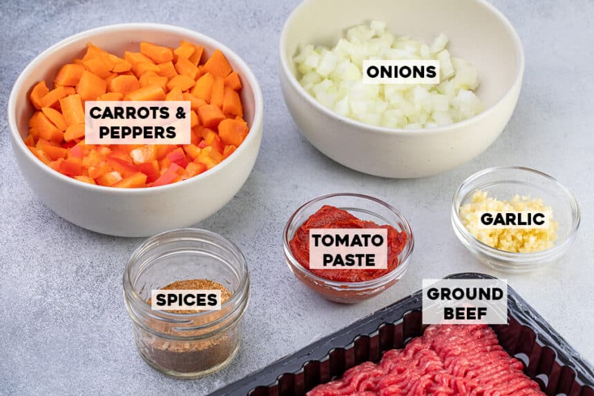 Chili ingredients to cook with titles of carrots & peppers, onions, spices, tomato paste, garlic, and ground beef in glass and ceramic bowls