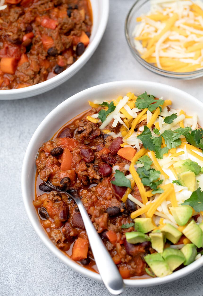 Red chili with black, deep red, orange ingredients, with green white and yellow toppings in bowl with spoon nearly overhead