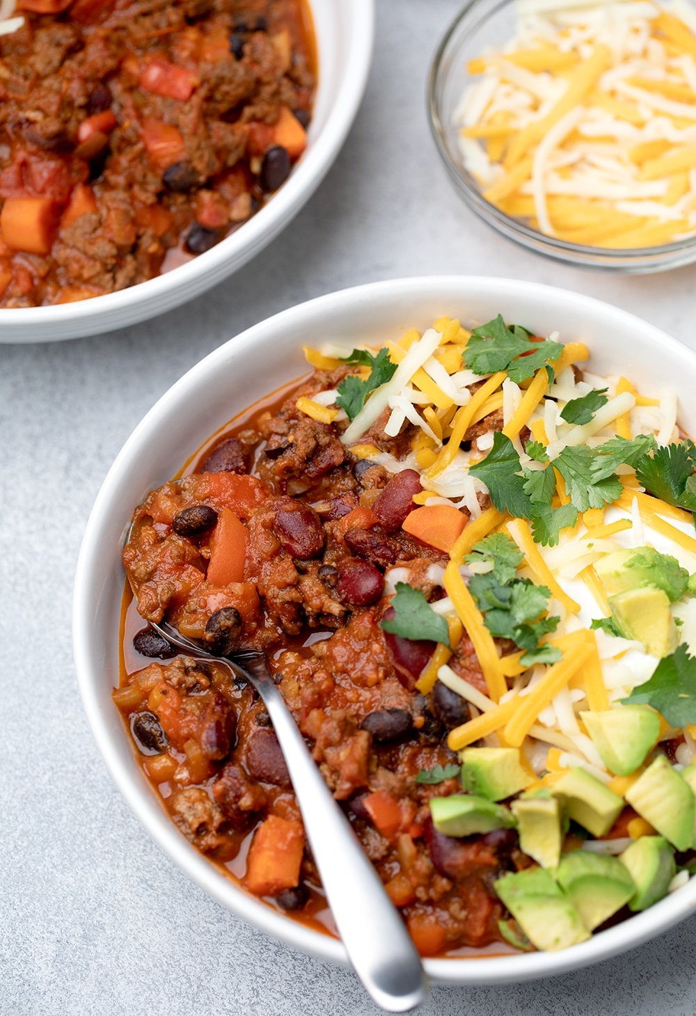 The chili served with green white and yellow toppings in bowl with spoon nearly overhead.