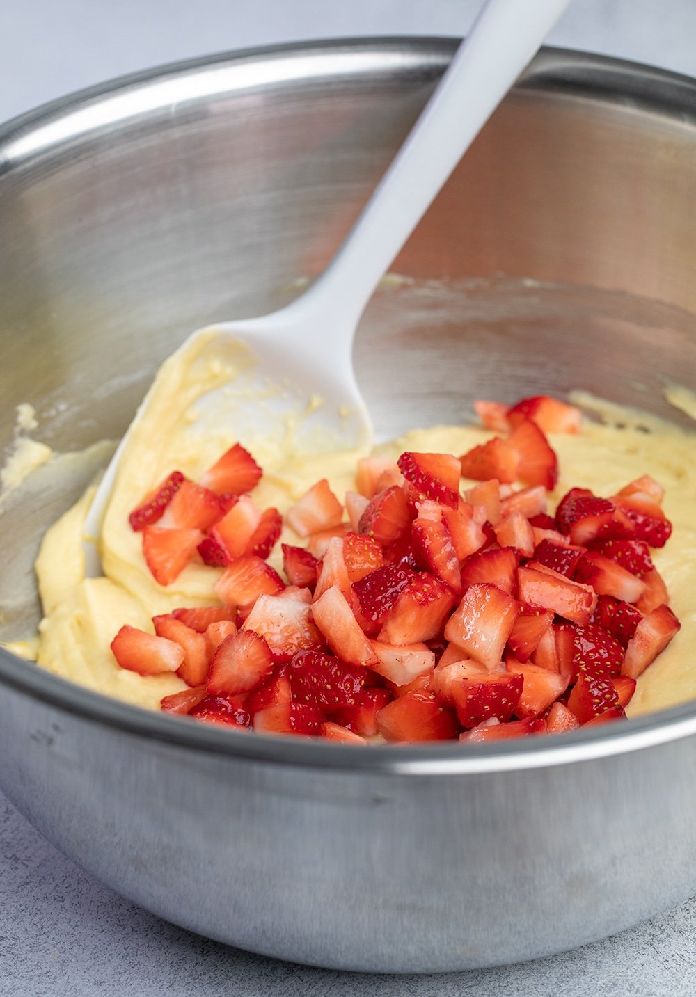 Chopped strawberries on yellow batter in metal bowl with spatula.
