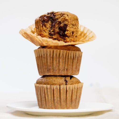 stack of 3 banana oatmeal muffins in brown paper liners on small white plate with top muffin broken in half