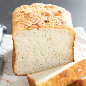 loaf of gluten free bread with a light brown crust, sesame seeds on top, and a few slices cut on white paper