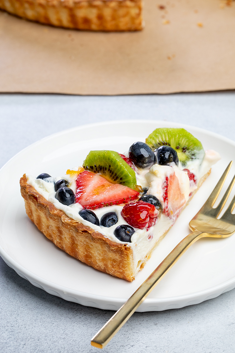 Slice of fruit tart with white filling mixed fruit on top in light brown shell with rest of fruit tart in background