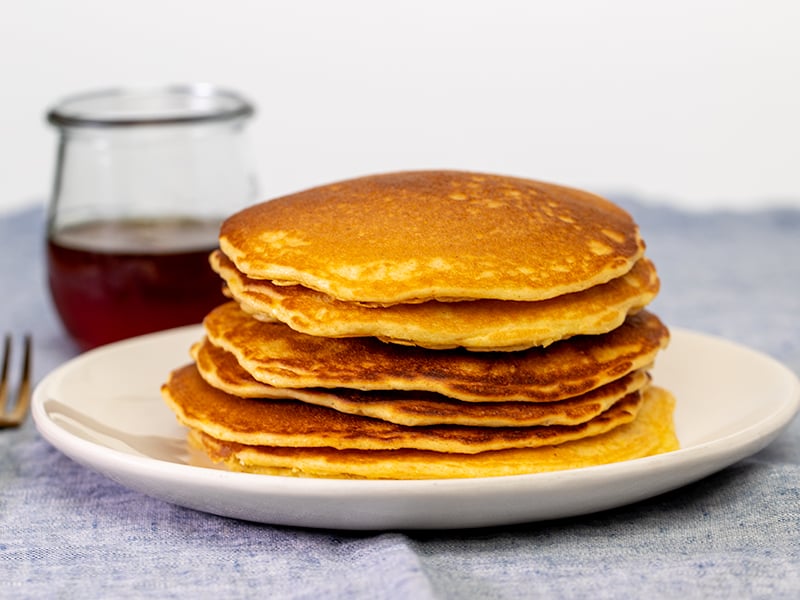 stack of 6 yellow pancakes on white plate on blue cloth