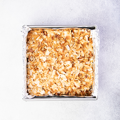 7 layer bars with toasted coconut chips on top in square metal baking pan