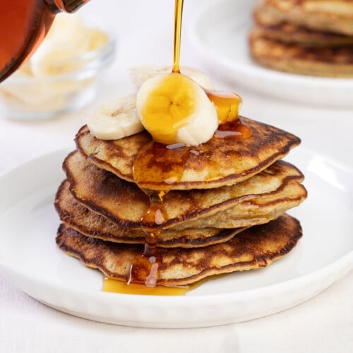 stack of 5 banana pancakes on white plate with sliced bananas on top and syrup bottle pouring