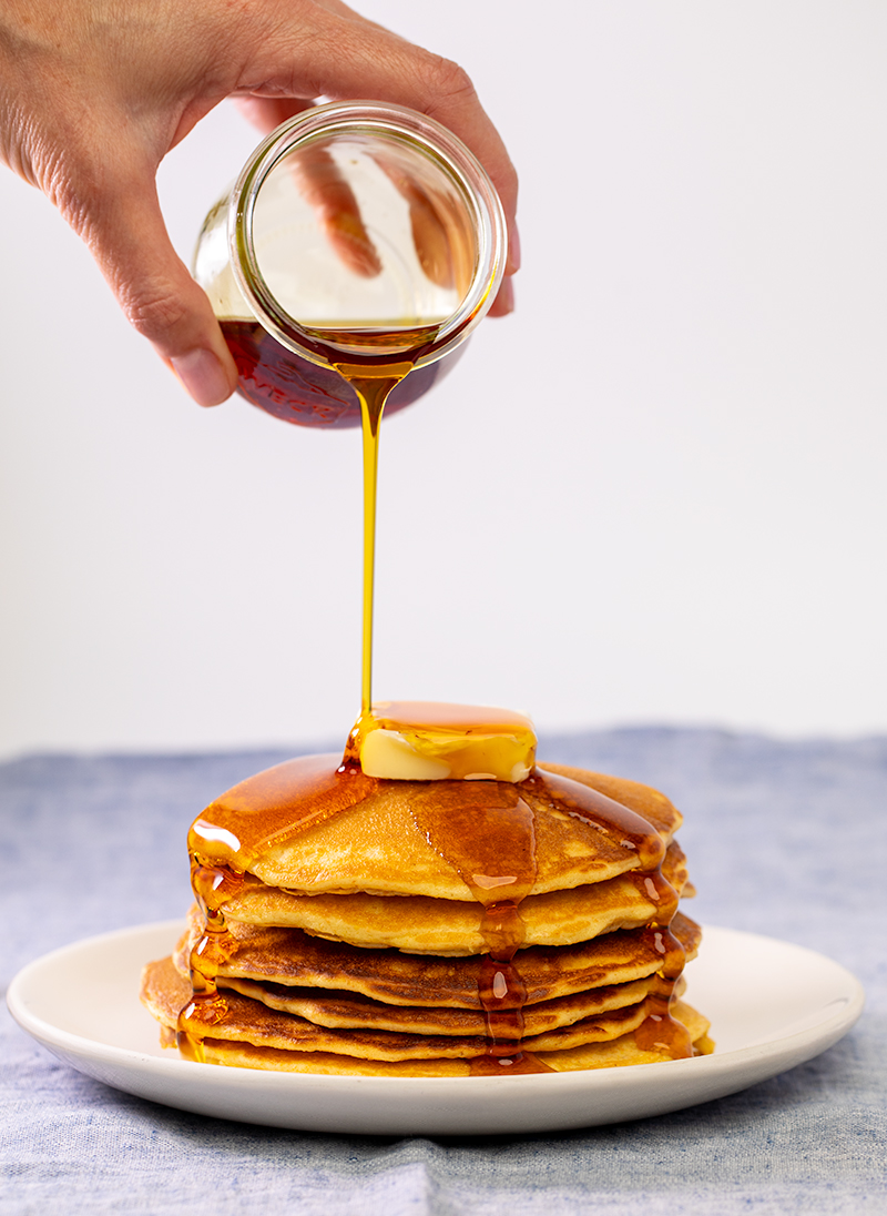 hand pouring syrup on stack of 6 cornmeal pancakes with pat of butter on white plate and blue cloth