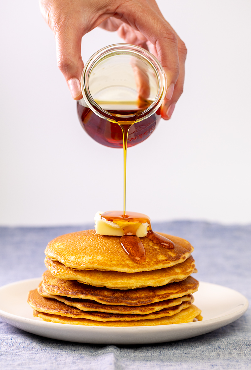 Maple syrup beginning to pour on stack of yellow cornmeal pancakes on white plate