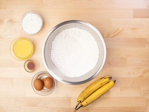 Dry ingredients for banana bread in metal mixing bowl with eggs, bananas, melted butter and sour cream in bowls