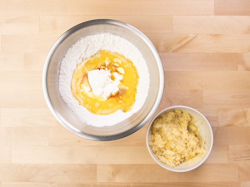 Dry ingredients in mixing bowl with sour cream, melted butter and eggs, with mashed bananas in small bowl