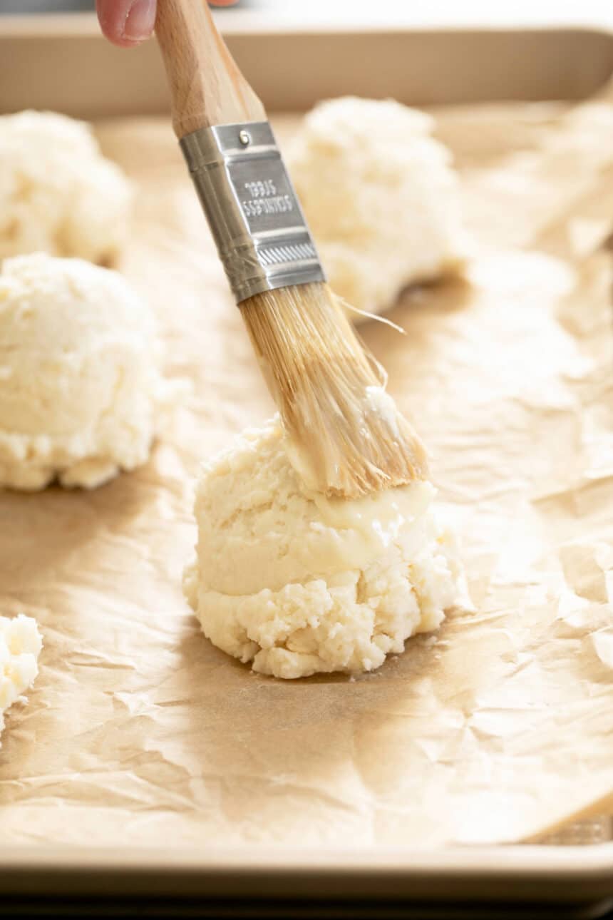 Raw biscuit being brushed with melted butter on brown paper.
