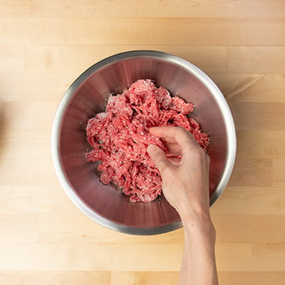 Hand breaking up beef and tossing with ground Parmesan cheese in metal round mixing bowl