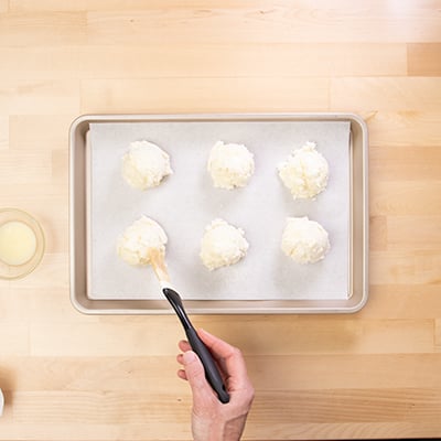 Hand brushing pastry brush with melted butter on raw drop biscuits on paper lined tray