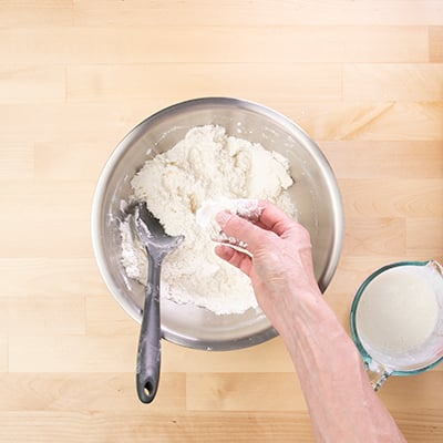 Hand holding flattened chunk of butter to make layered biscuits over bowl of dry ingredients