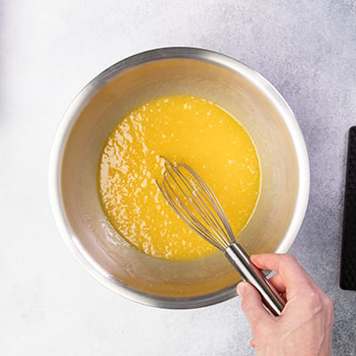 Hand holding whisk in metal mixing bowl with yellow mixture