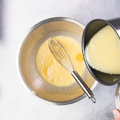 Hand pouring melted butter and sugar into whisk in bowl with beaten eggs