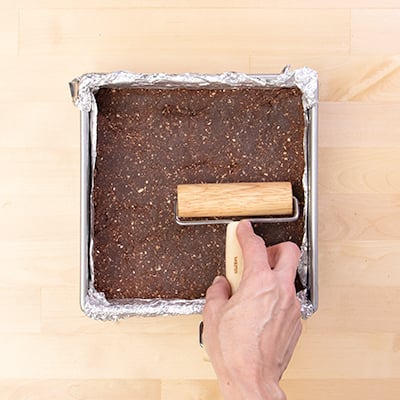 Hand pressing small handheld roller on protein bar mixture in metal baking pan