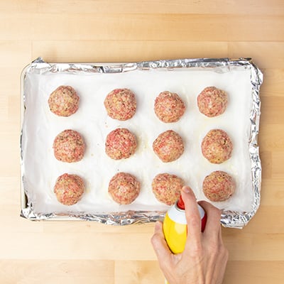 Hand spraying cooking oil spray on raw shaped meatballs on paper lined baking tray