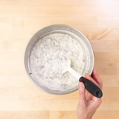 Hand stirring spatula mixing in Oreo cookie crumbs to cheesecake filling