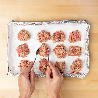 Hands holding two spoons placing portions of meatball mixture on paper lined baking tray