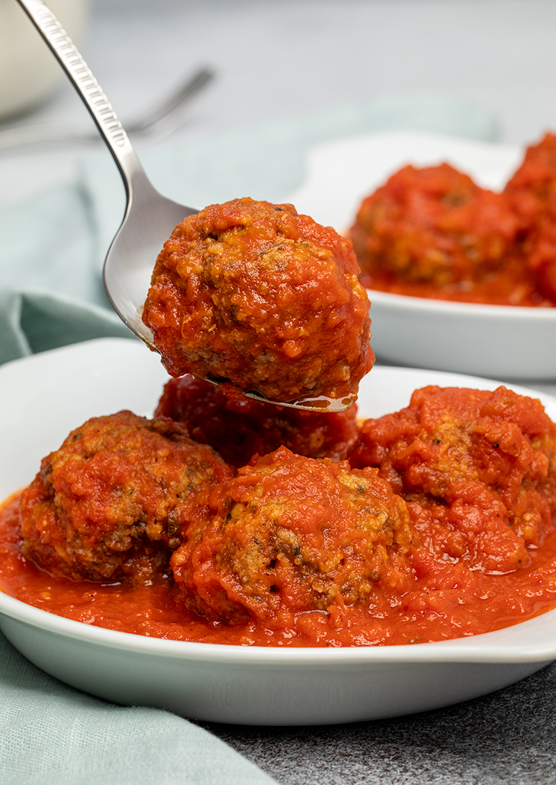 Meatball on metal serving spoon being placed on top of 4 other meatballs with tomato sauce in white dish