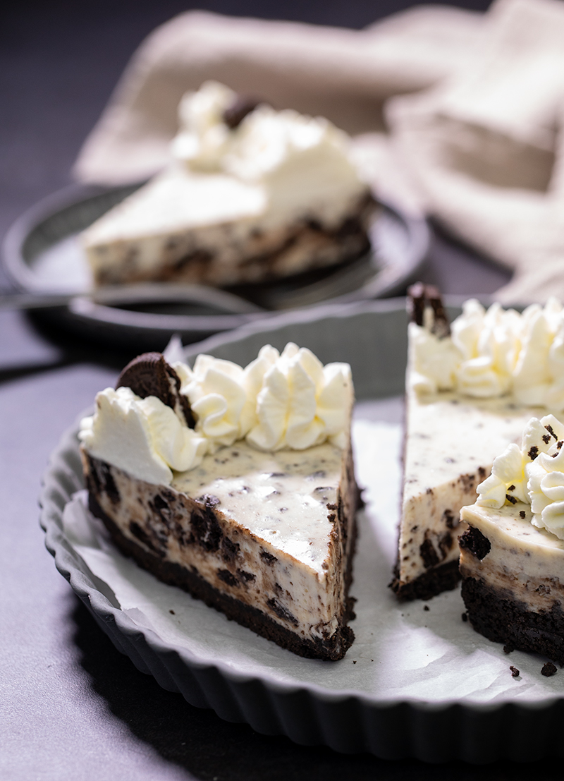 No bake Oreo cheesecake on white paper in tart pan with slice in background on small gray plate with fork