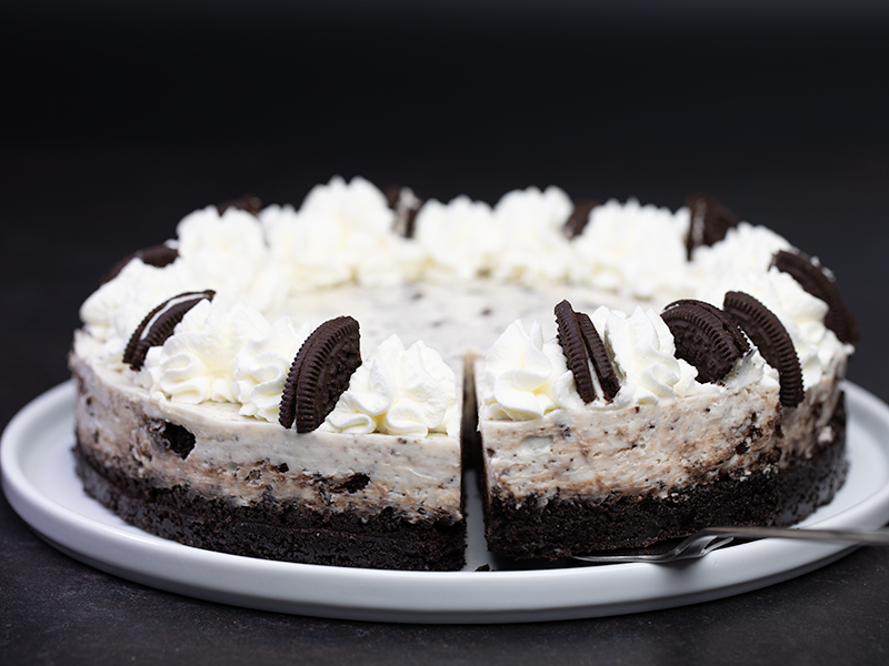 No bake oreo cheesecake with whipped cream and cookie decoration on white serving platter with one slice cut