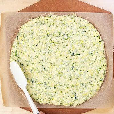 White spatula shaping dough for zucchini pizza into flat round on brown paper