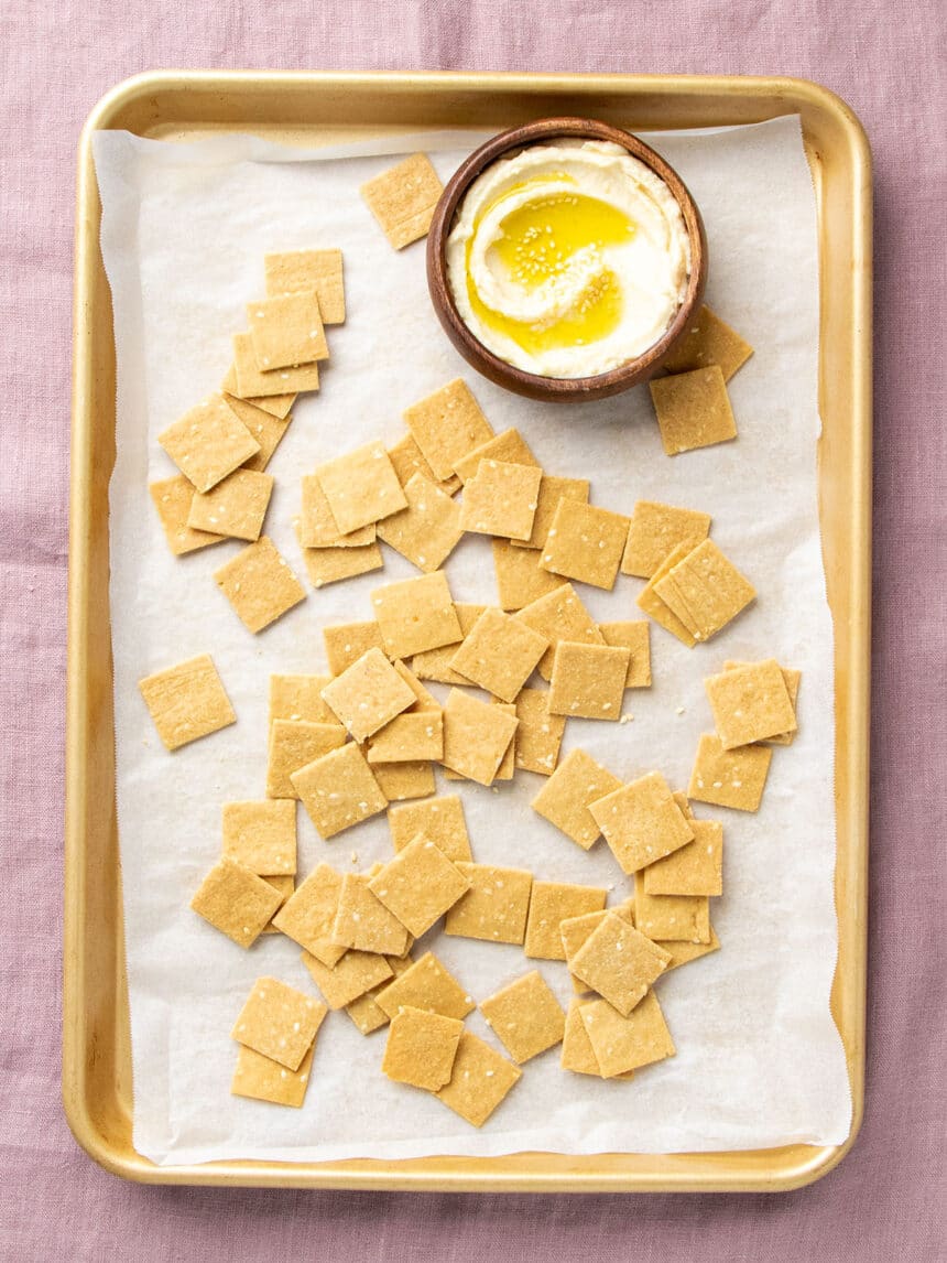 light brown square crackers with sesame seeds on white paper on baking tray with hummus