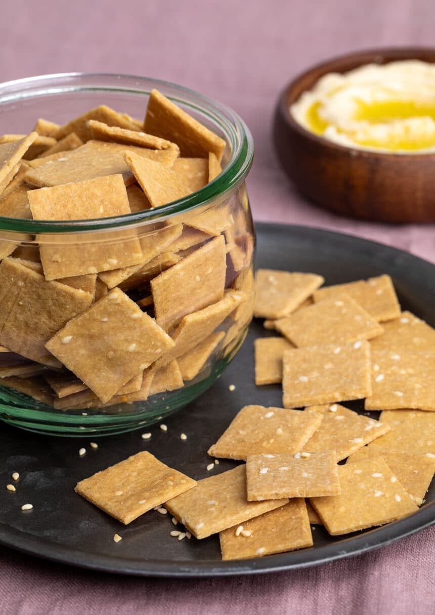 square almond flour crackers with sesame seeds on black plate with more crackers in glass mason jar