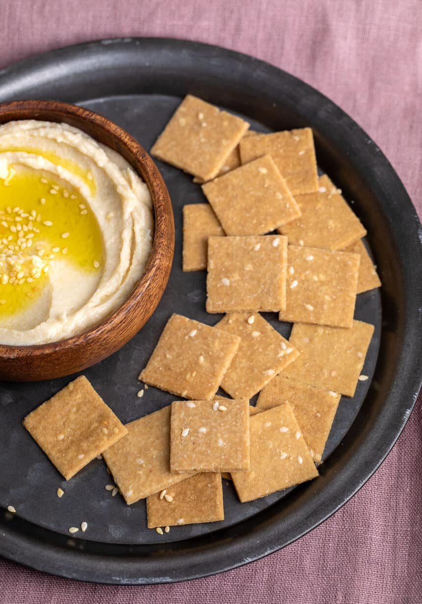 square brown crackers with sesame seeds on black round plate with hummus in small round wood bowl