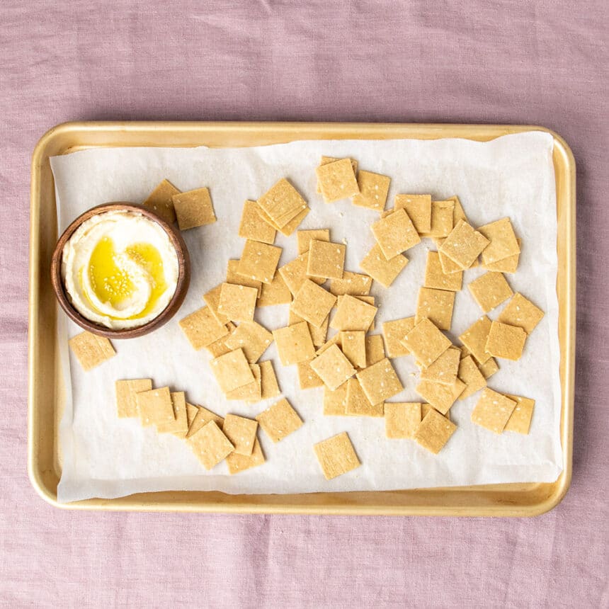 light brown baked 1-inch square crackers on white paper on baking tray with olive oil and hummus in small wood bowl