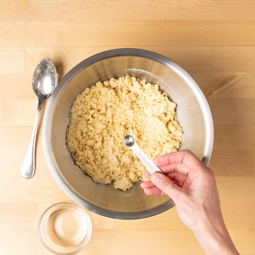 crumbly light brown dough in mixing bowl with hand holding 1/4-teaspoon over bowl