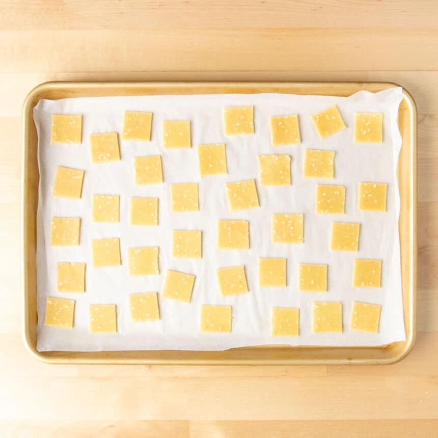 1-inch light brown raw cracker squares on white paper on golden baking tray