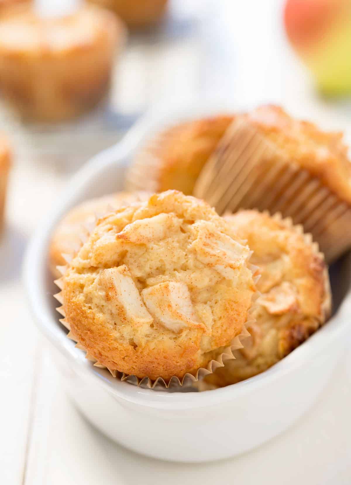 White bowl with light brown apple muffins in brown papers with front apple on its side