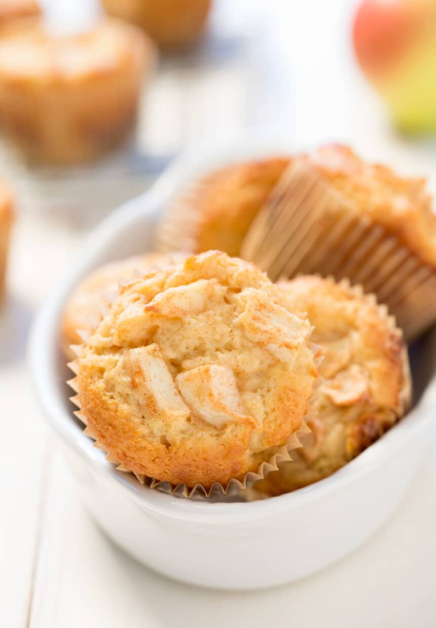 White bowl with light brown apple muffins in brown papers with front apple on its side