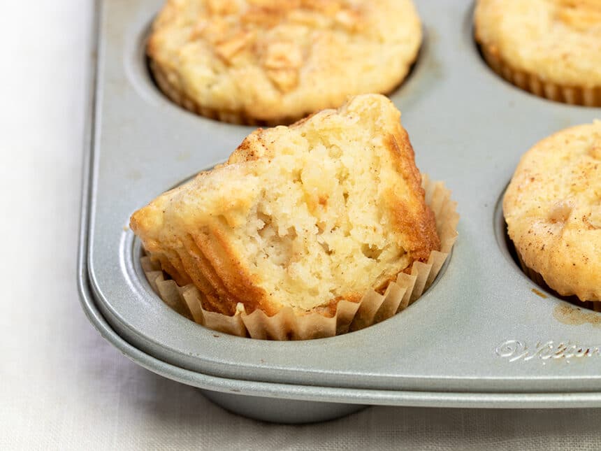 Metal muffin tin with light brown muffins with front muffin broken to reveal inside